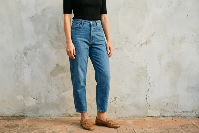 Um top de malha canelada preta por dentro de jeans cocoon azul médio, combinado com mocassins de couro cor tan mostrando o tornozelo, contra uma parede cinza claro texturizada.
