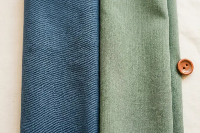Close-up de tecido de scrub de gabardine azul com trama fina ao lado de tecido de scrub de poliviscose verde macio, realçando suas texturas e tramas distintas em uma superfície branca.