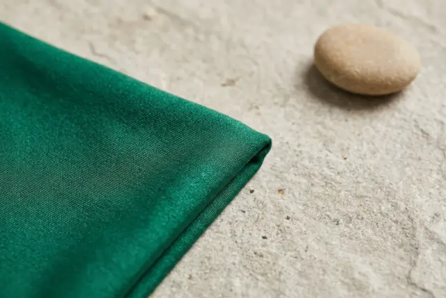 Um close-up macro de tecido de maiô de nylon-lycra verde esmeralda macio e perfeitamente preservado, mostrando sua cor vibrante e textura suave, cuidadosamente disposto sobre uma superfície de pedra c