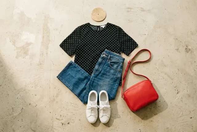 Sobre uma superfície de concreto claro, uma blusa preta de bolinhas, calça jeans azul de perna reta, tênis brancos de couro e uma bolsa transversal de couro vermelho vibrante.