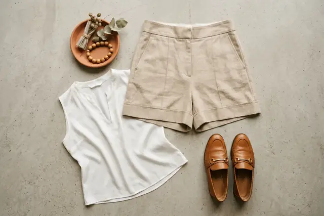 Shorts de linho bege sob medida, precisamente dobrados para revelar a barra pesada e vincada, com blusa de seda branca sem mangas e mocassins de couro caramelo, sobre uma superfície de concreto cinza