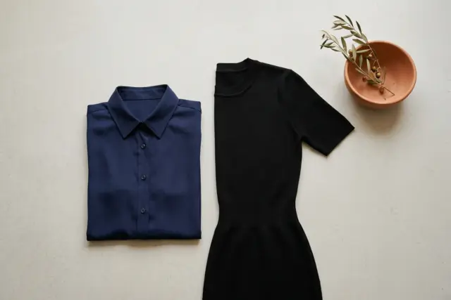 Disposição plana de uma camisa de poliéster azul marinho perfeitamente lisa e um elegante vestido de tricô preto com elastano, ambos sem vincos em um chão de estúdio cinza claro e limpo.