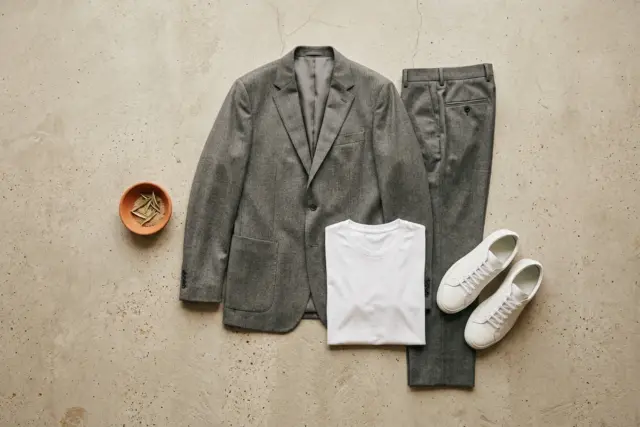 Disposição plana de um blazer de lã fria cinza e sua calça combinando, uma camiseta de algodão branca e tênis de couro brancos, dispostos em um piso de concreto claro.