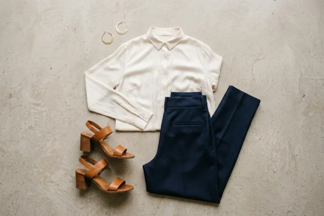 Blusa de seda off-white e calça de lã de alfaiataria azul-marinho, dispostas ao lado de sandálias de salto bloco de couro caramelo e brincos delicados de metal dourado, em concreto cinza-claro.