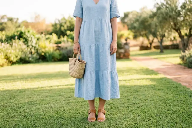 Convidada vestindo vestido midi de linho azul claro, sandálias de salto bloco caramelo e uma bolsa de palha pequena, em um gramado macio e ensolarado.